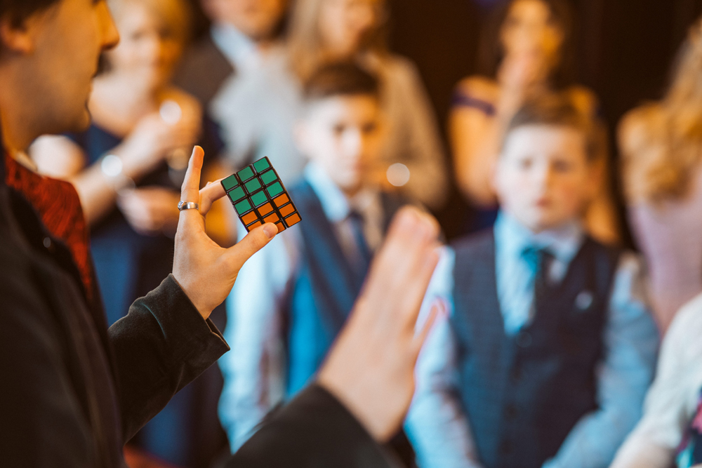 A close up of a magician holding a Rubick's cube, with wedding guests in the background