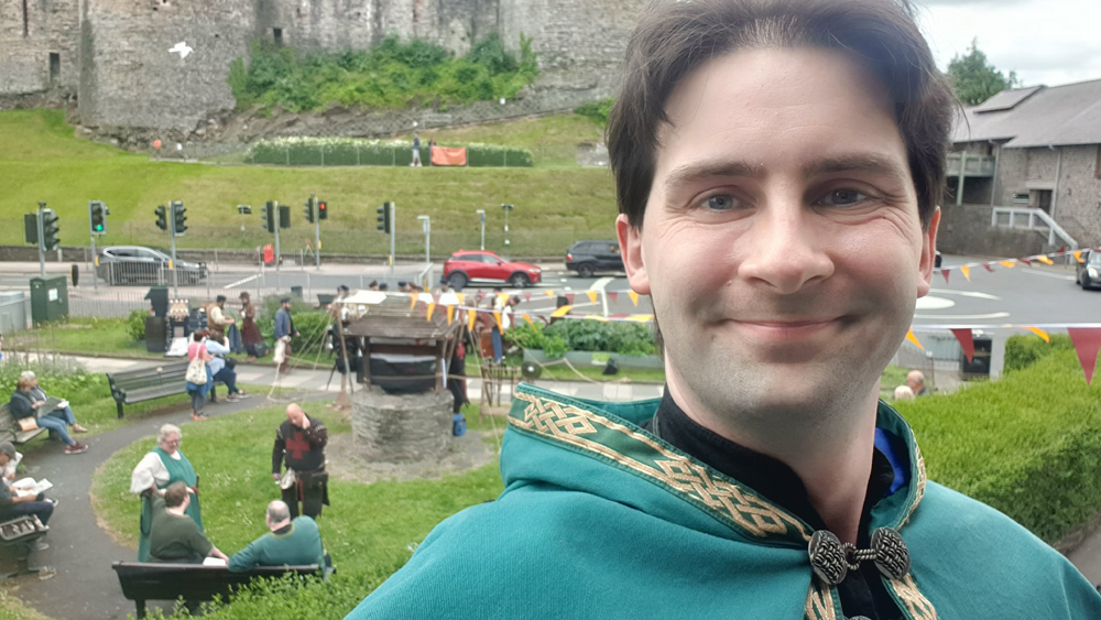 A selfie of Jay Gatling, the wizard of Conwy. He is dressed in wizard robes of green and gold. In the background is Conwy castle, and people dressed as knights.