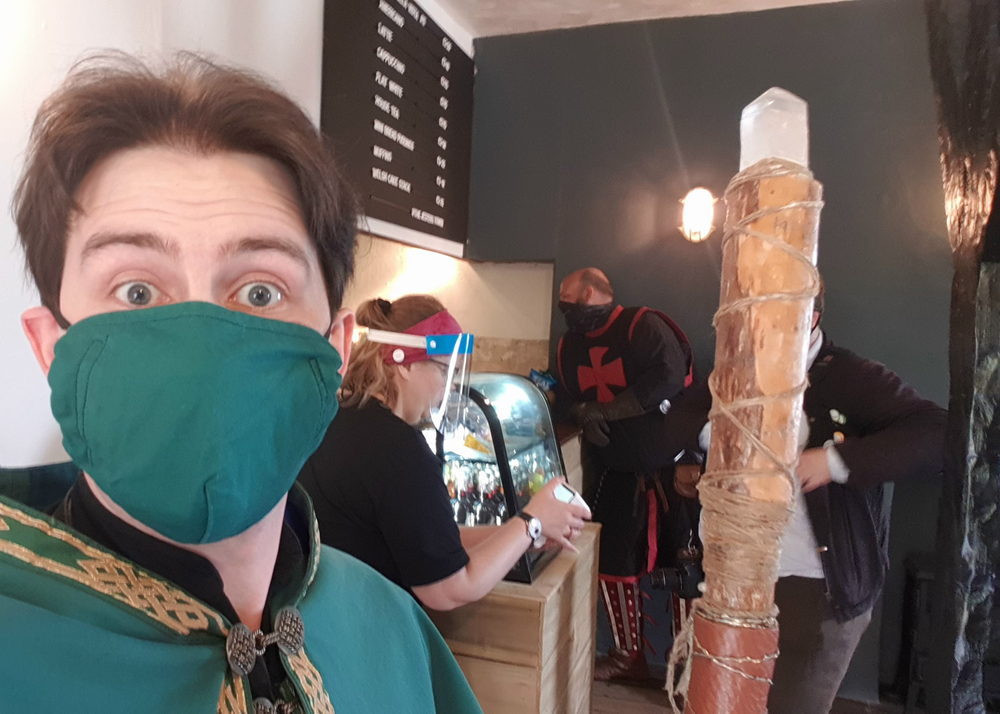A selfie of Jay Gatling, the wizard of Conwy. He is dressed in wizard robes of green and gold, and holds a wizard's staff. He is inside the Jester's Tower coffee shop, and in the background is a person serving customers at the counter. One customer is dressed as a knight.