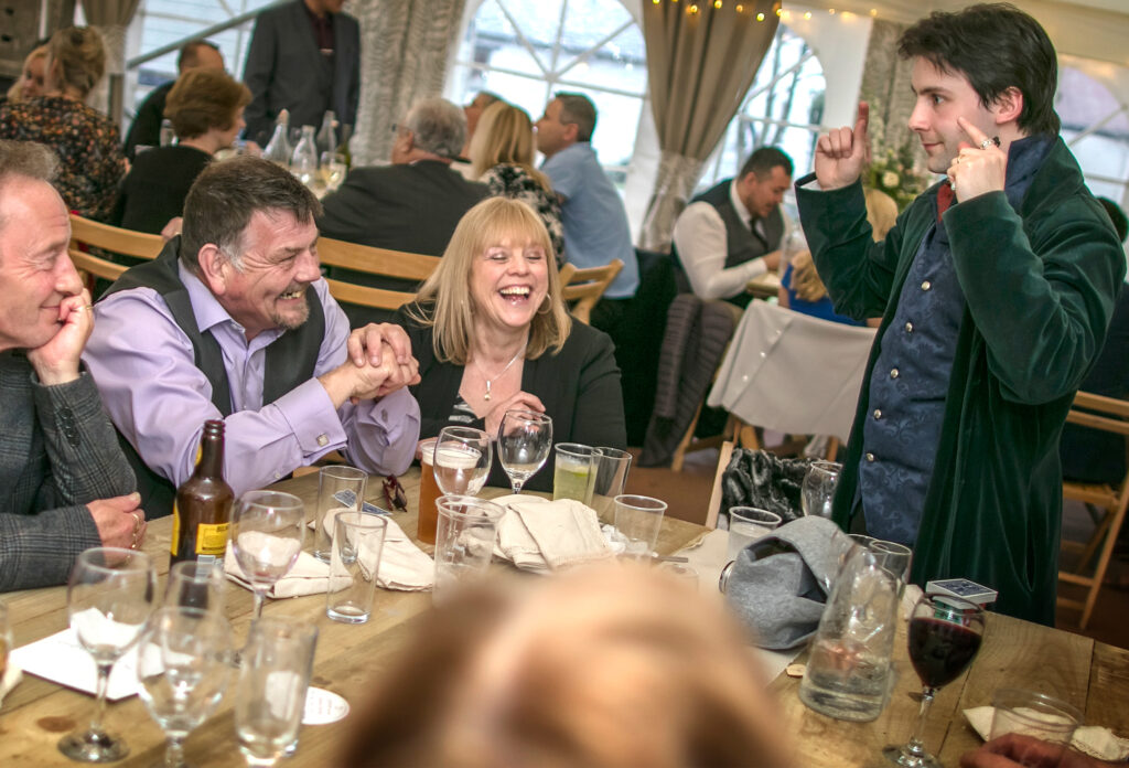 A table of guests at a wedding. They are laughing and smiling at the magician who is showing them a trick.