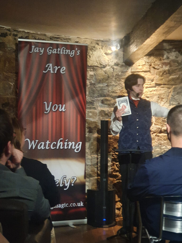 Magician Jay Gatling holds a jumbo deck of playing cards. He is standing in front of a banner with the words "Are You Watching Closely?" There are stone walls and wooden beams in the background.