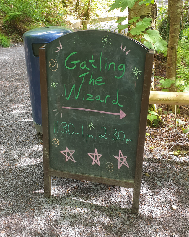 A wooden blackboard sign on a gravel path in a forest. The text reads "Gatling the Wizard 11.30am, 1pm, 2.20pm"