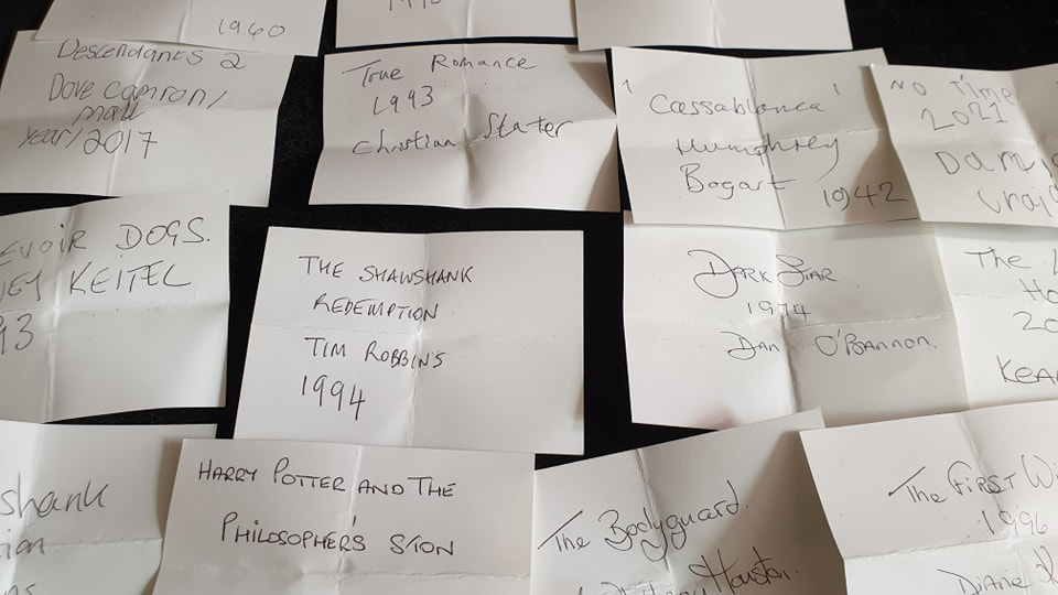 Small pieces of paper laid out on a black table. Each has the name of a film written on it, along with the year, and the main actor. There are around 15 films, each in different handwriting.