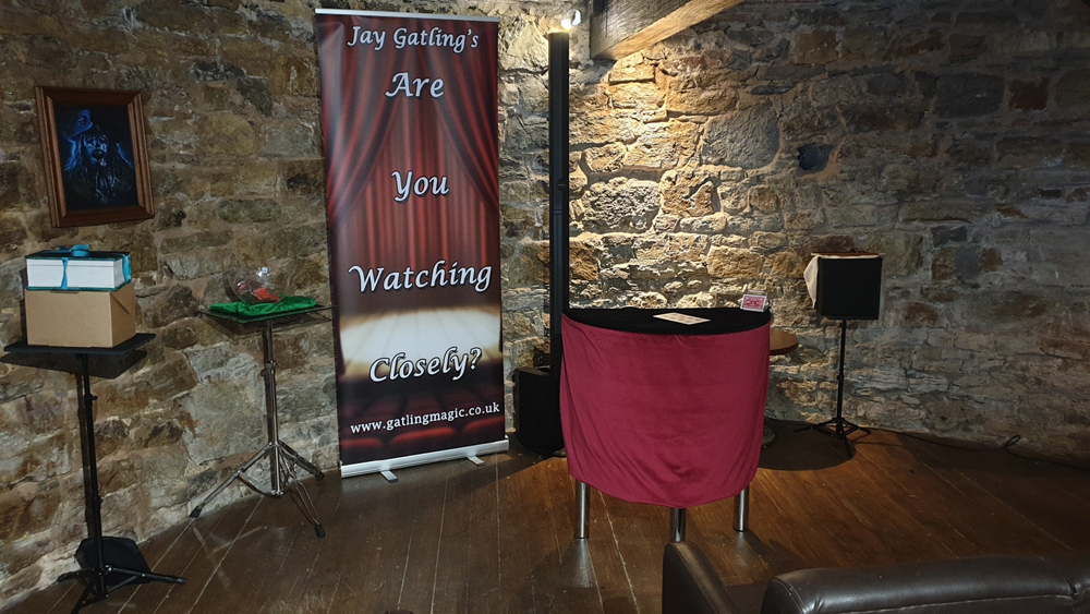 Three small tables piled high with magical props. A fourth, larger table stands in front, with a red velvet drape. The room has a curved stone wall, and a wooden beam can be seen across the ceiling.