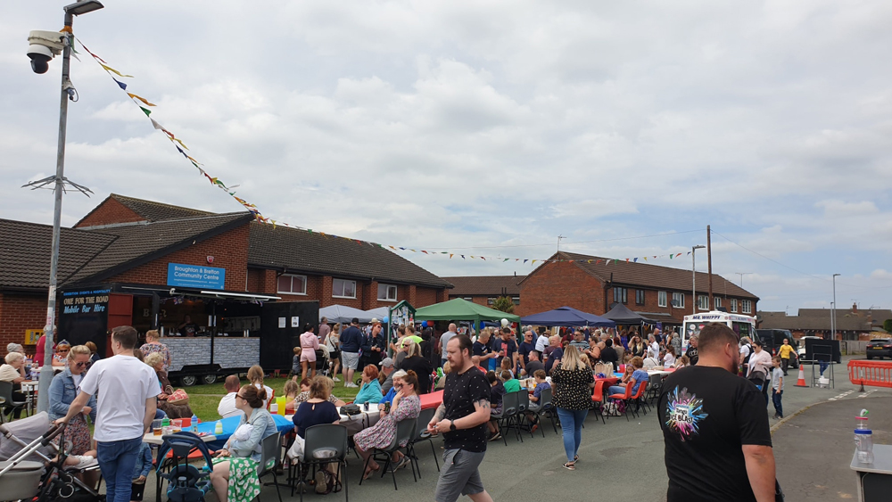 A street party. There are tables laid end to end in the road, with people sat at chairs. Broughton and Bretton Community Centre is in the background.