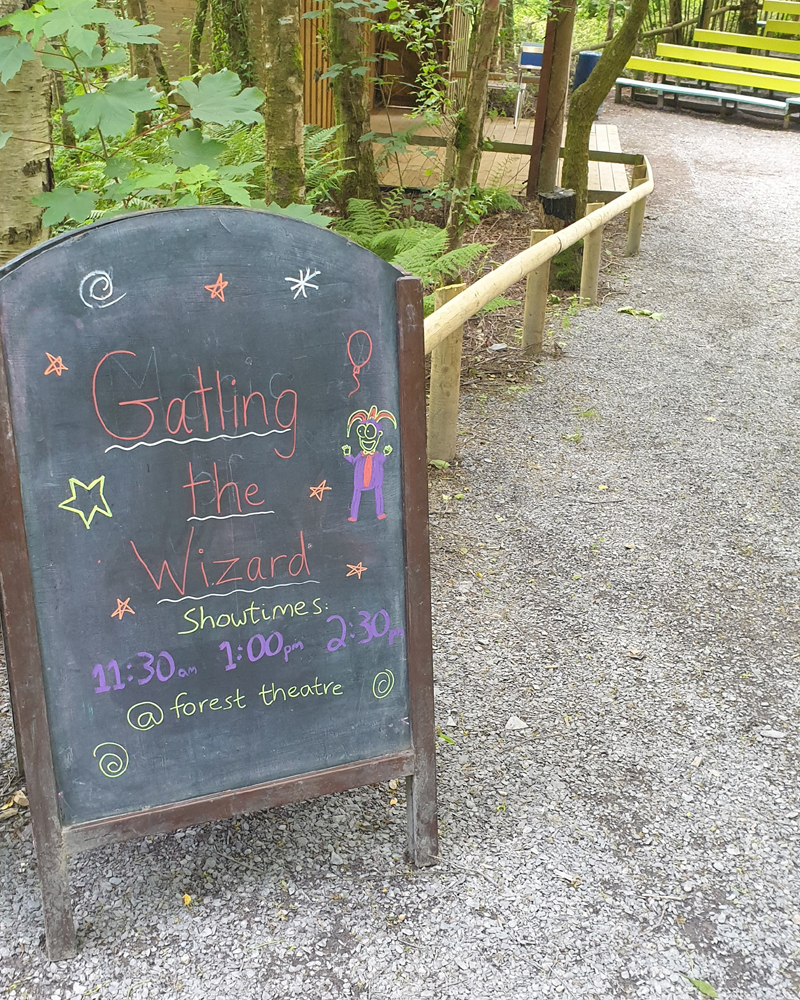 A wooden blackboard sign on a gravel path in a forest. The text reads "Gatling the Wizard showtimes 11.30am, 1pm, 2.20pm"