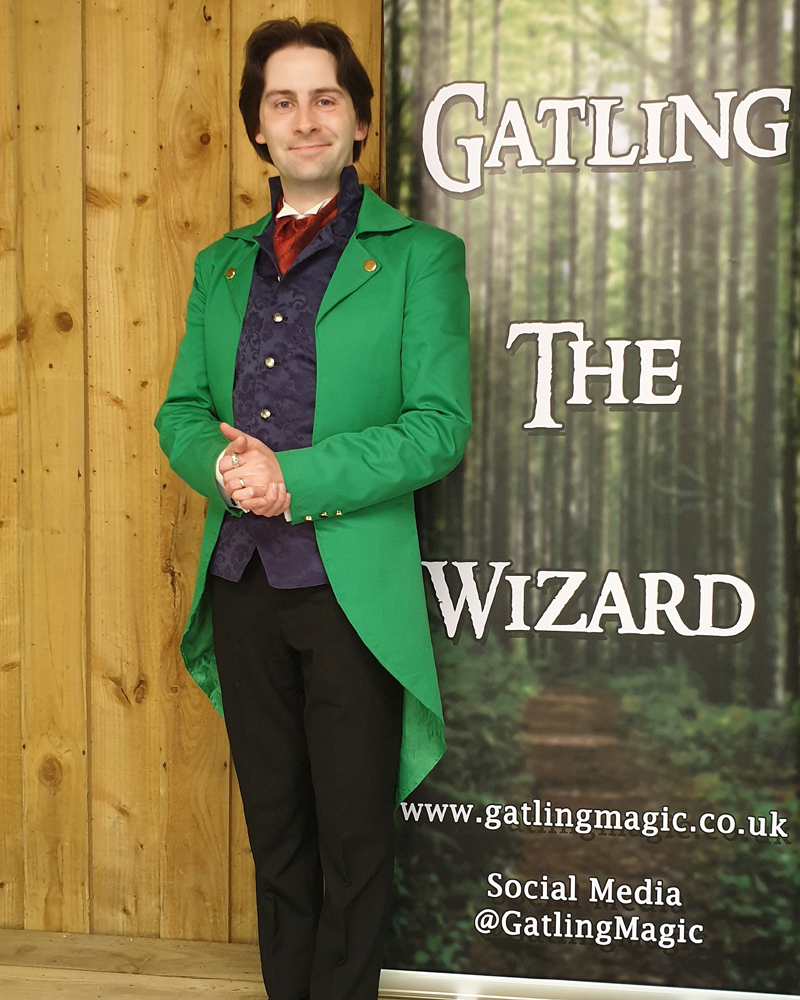 Jay Gatling, smiles at the camera. He has dark hair, and is wearing a bright green tailcoat, with trousers and a smart waistcoat. Behind him is a banner that reads "Gatling the Wizard".