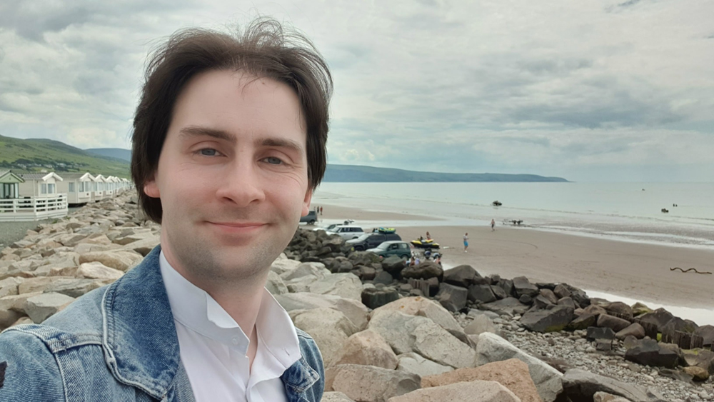 Jay Gatling smiles at the camera. He has dark hair, and is wearing a blue denim jacket over a white open-necked shirt. He is standing on a rocky section of shore. Behind him is the beach, and the sea.