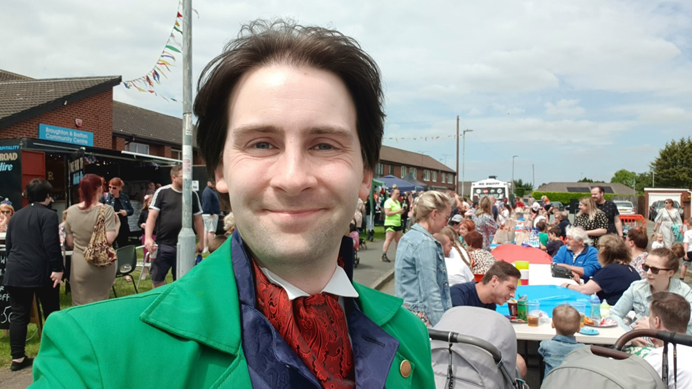 Jay Gatling standing in front of Broughton and Bretton Community Centre. There are tables in the road, with people sat at them, eating and drinking.