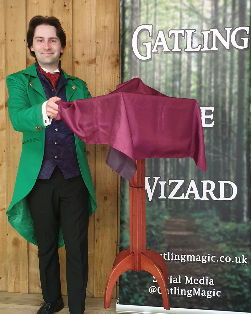 Jay Gatling, smiles at the camera. A wooden table is floating in the air in front of him. He has dark hair, and is wearing a bright green tailcoat, with trousers and a smart waistcoat. Behind him is a banner that reads "Gatling the Wizard".