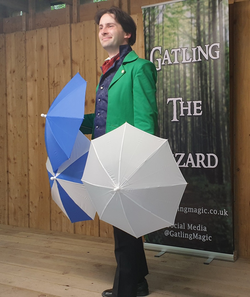 Jay Gatling holds two parasols, one white, one blue and white. Behind him is a banner that reads "Gatling the Wizard".