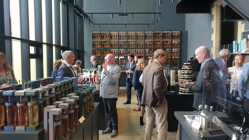 Smartly dressed people in a room with grey walls. There are displays of whisky botles, and a stand with multiple tiers of cupcakes on it,