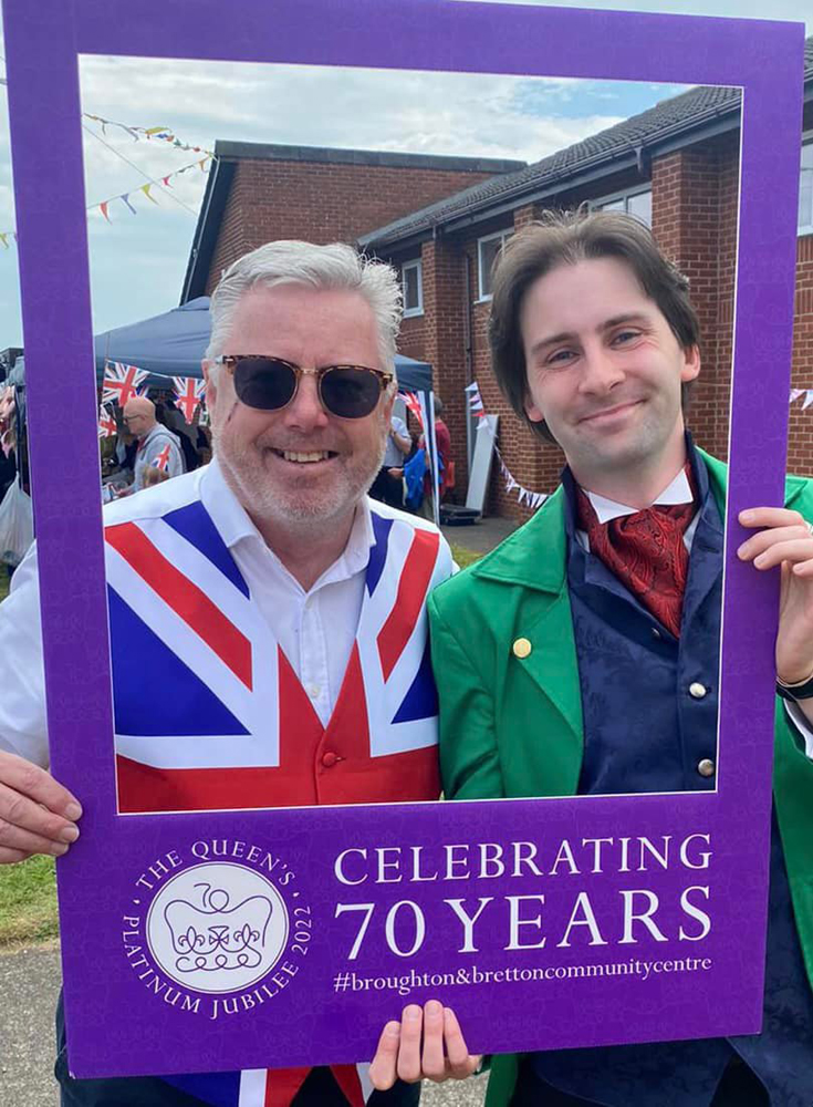 Two people are holding a purple "selfie-frame" that reads "Celebrating 70 years". The man on the left is wearing a Union Flag waistcoat. Jay Gatling is standing to the right, wearing a bright green tailcoat, a blue waistcoat, and a red cravat.