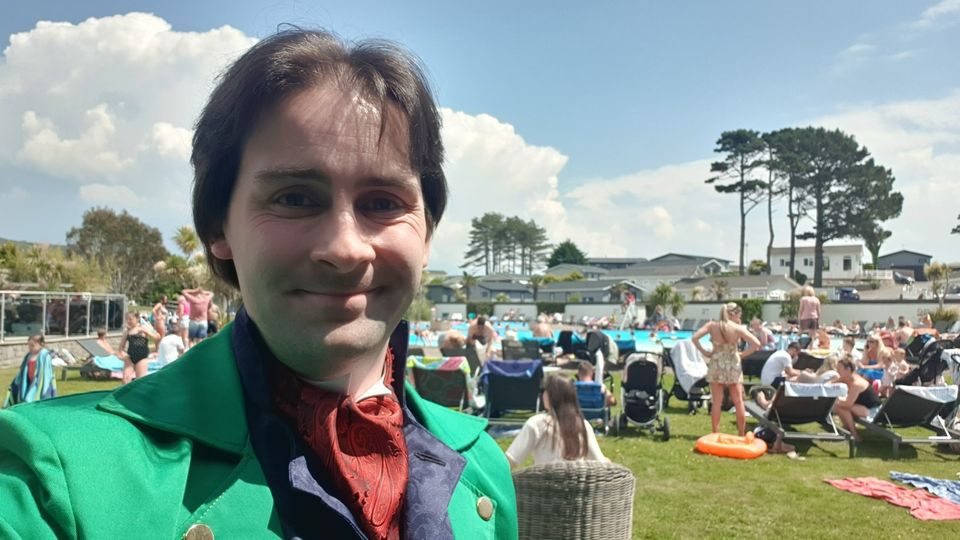 Jay Gatling is standing in front of a crowd of people who are gathered around an outdoor pool. The sun is shining, and there are fluffy white clouds in the sky.