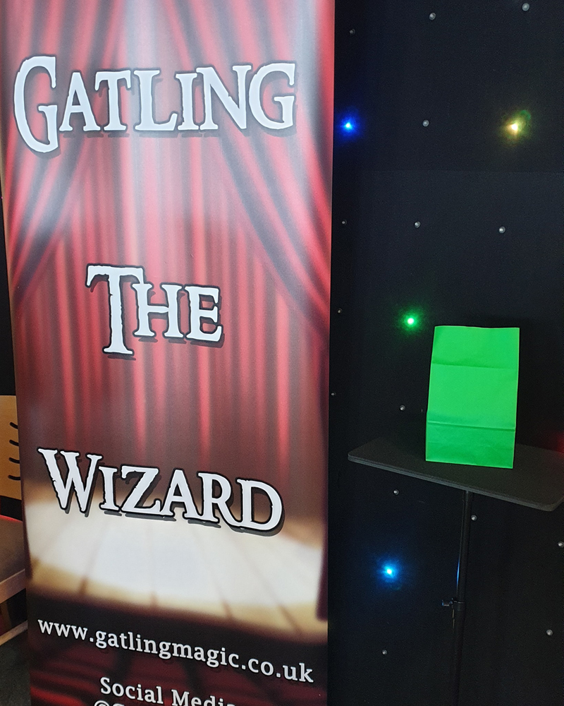 A red banner reads "Gatling the Wizard". Next to it is a small table with a bright green paper bag on it. Behind both is a black cloth with lots of little bright lights on it.