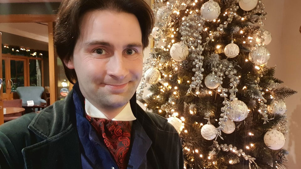 A dark haired man wearing a green velvet frock coat, blue waistcoat, and red cravat stands in front of a Christmas tree.