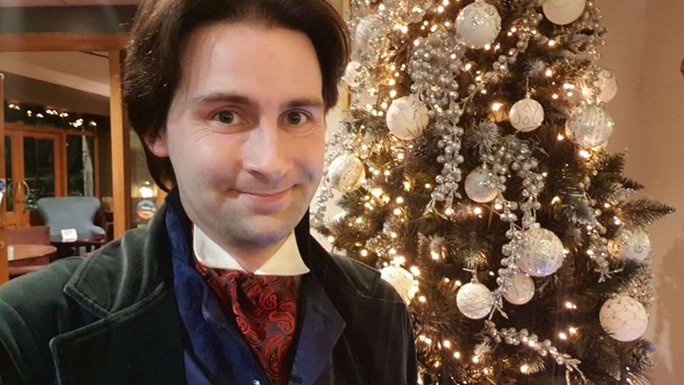 A dark haired man wearing a green velvet frock coat, blue waistcoat, and red cravat stands in front of a Christmas tree.